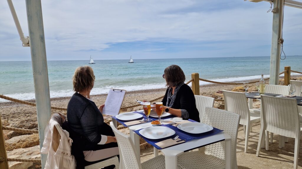 Dos mujeres sentadas en una mesa frente al mar, disfrutando de la vista y leyendo el menú. En el horizonte, se pueden ver veleros navegando.