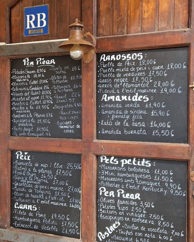20240405_120952 Puerta de madera con varios menús escritos en pizarras negras. El menú incluye platos como patatas bravas, paella de marisco, pescado, carnes y postres. Hay una luz de pared y un letrero azul con las iniciales "RB".