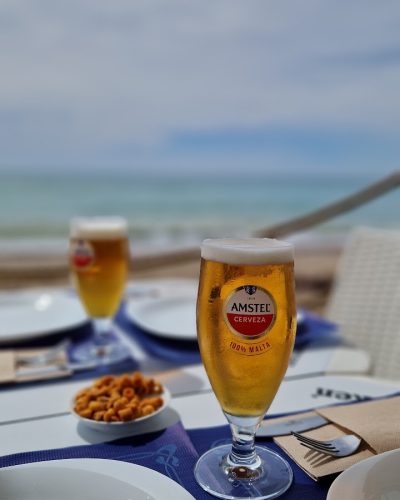 20240405_123005 Primer plano de una mesa con dos cervezas Amstel servidas en copas, un pequeño plato de snacks y platos vacíos, con el mar de fondo.